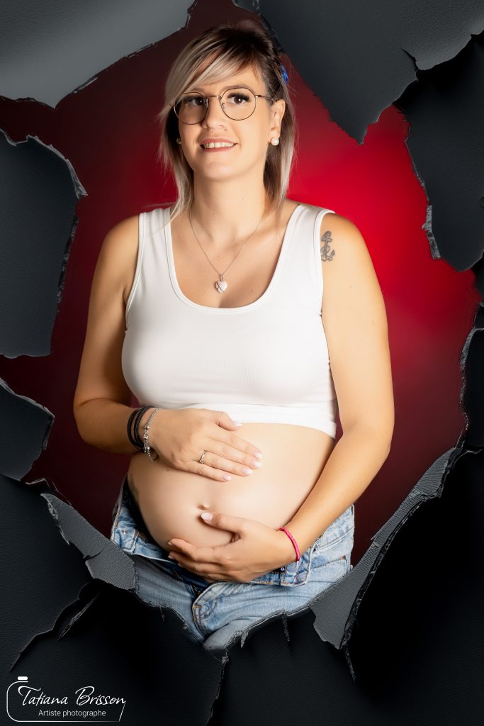 Femme enceinte souriante tenant son ventre rond devant un décor rouge avec effet papier déchiré à Mâcon/Tatiana Brisson photographe.